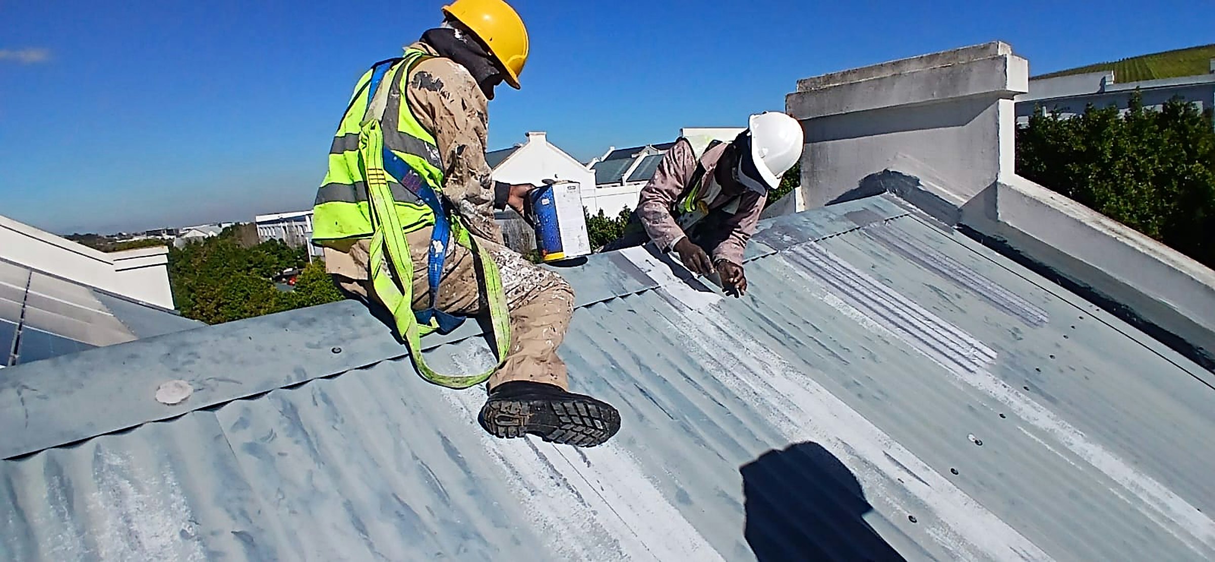 Two Neil Ambrose roofers in safety harnesses coating a metal roof under a clear Cape Town sky